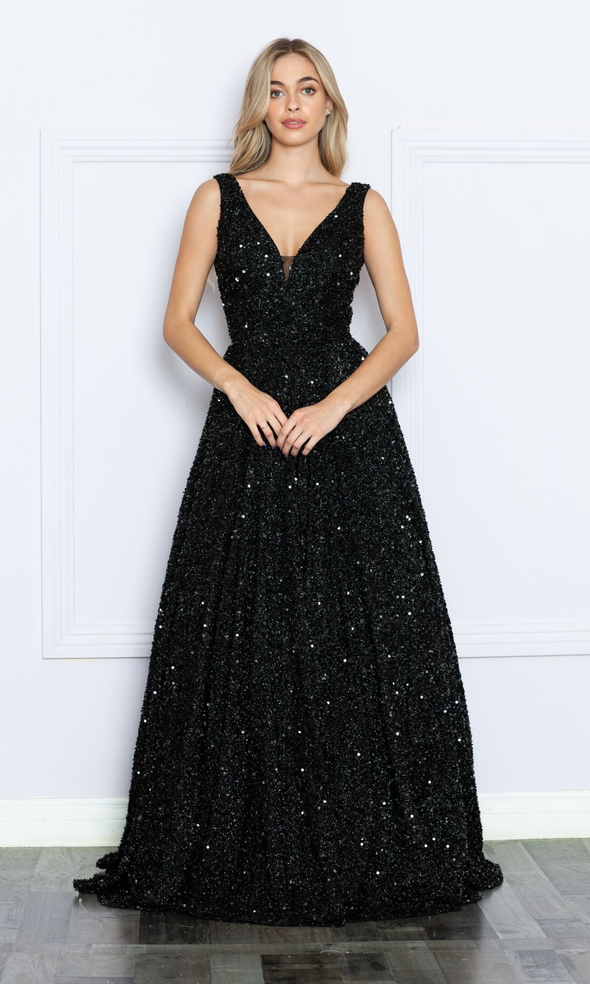 A woman stands against a white wall wearing the Long Black Sequin A-Line Prom Dress 9168, featuring a sleeveless, floor-length design with a deep V-neckline.